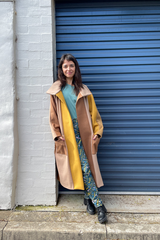 woman with long brown hair, wearing a camel and yellow patchwork wool felt coat, teal t-shirt, print pants and black boots with her hands in her pockets against a white brick wall