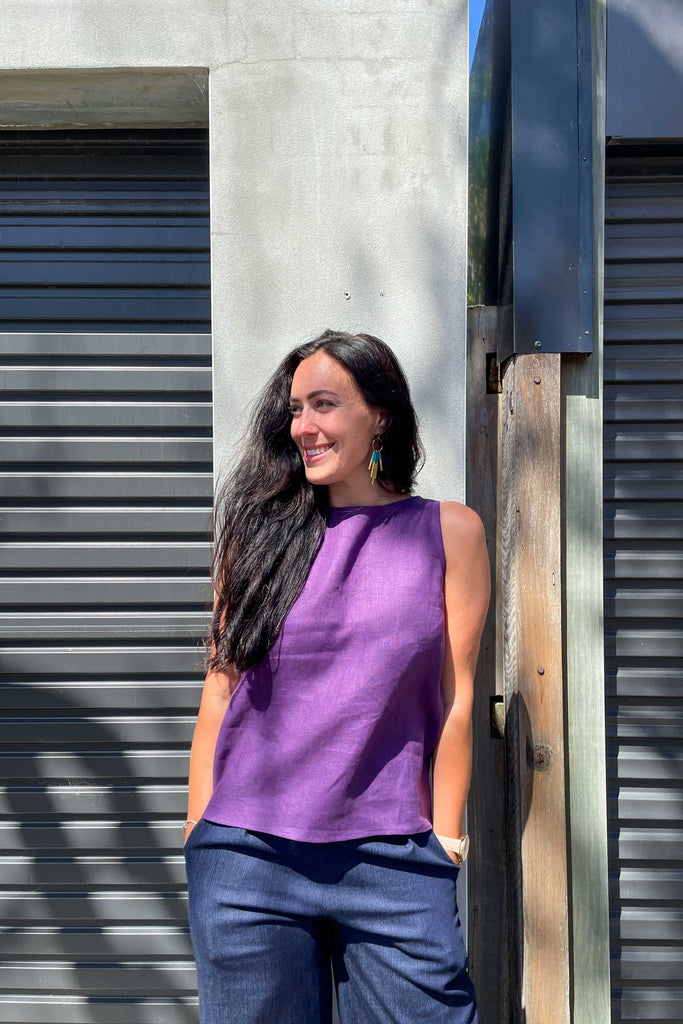 woman wearing violet purple linen summer shell top from earn and learn with denim culottes leaning against concrete wall